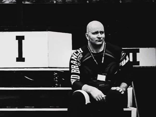 John Danaher seated beside a competition podium in a rashguard